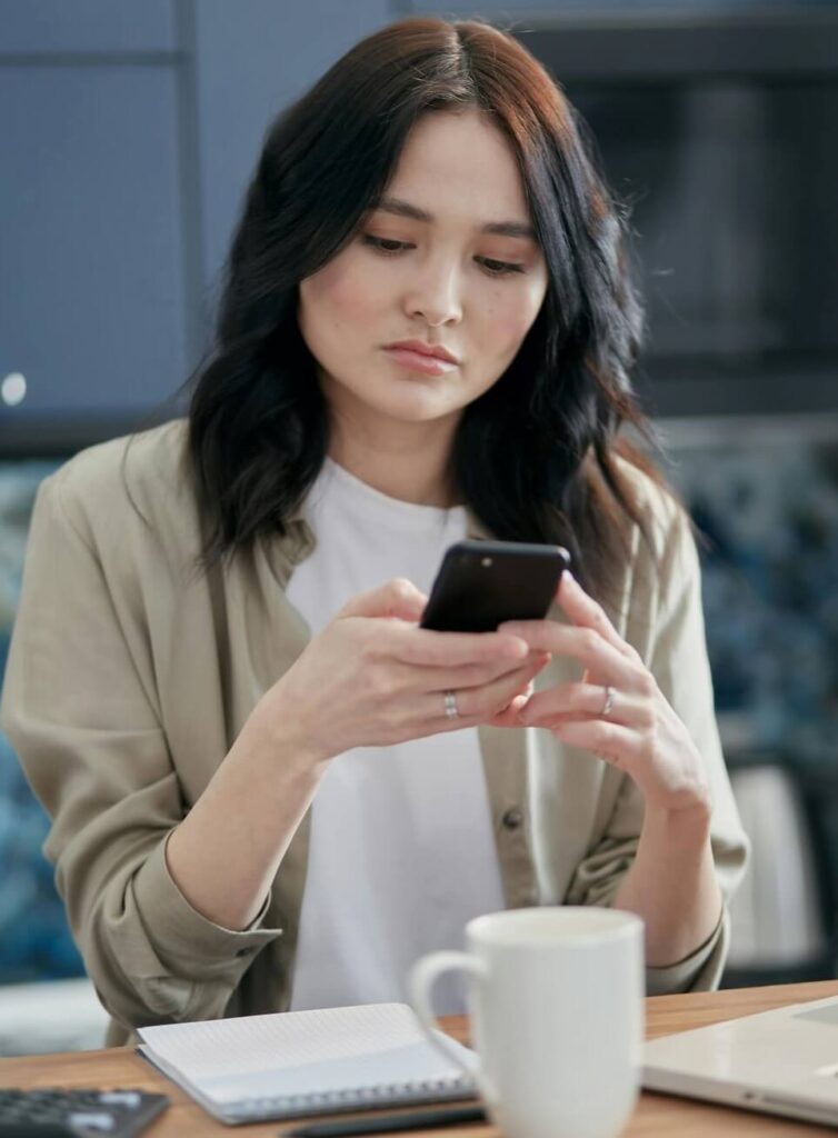 Mulher olhando o celular enquanto organiza suas finanças em casa com caderno, calculadora e café sobre a mesa