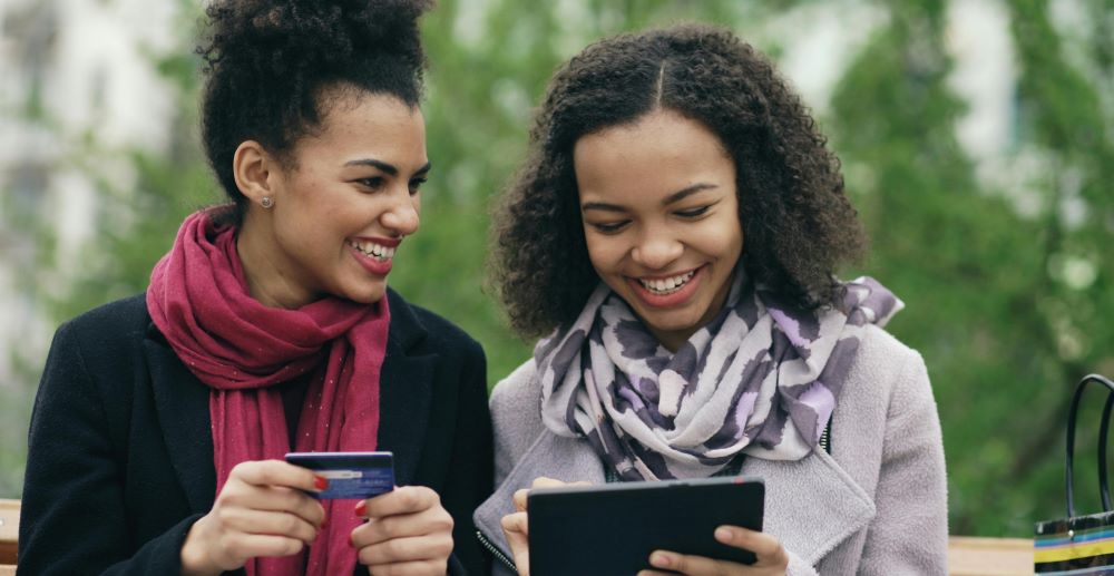 Duas mulheres consultando informações financeiras em um tablet, enquanto uma delas segura um cartão de crédito.