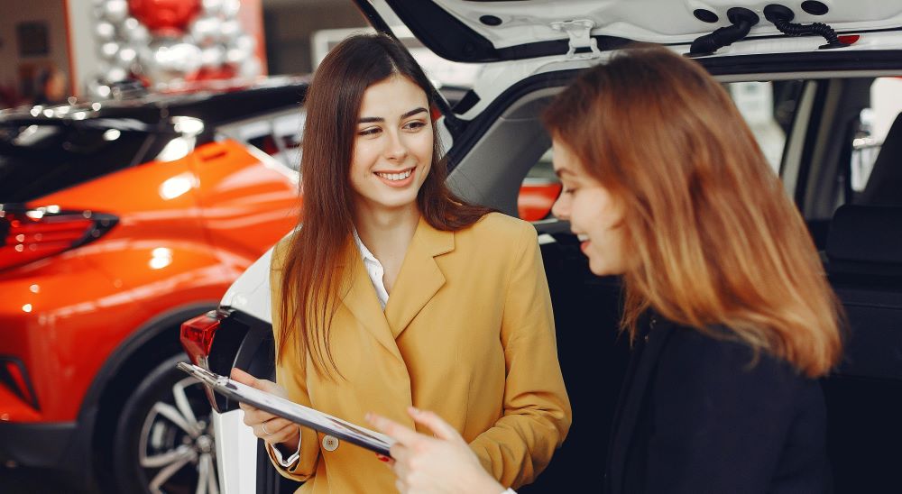 Duas mulheres analisando documentos ao lado de um carro em uma concessionária, representando planejamento financeiro e tomada de decisão para a compra de um veículo.