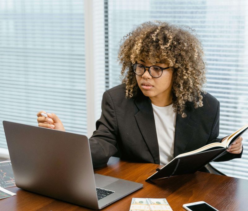 Mulher sentada em uma mesa de escritório analisando informações no notebook enquanto consulta um caderno, com dinheiro e celular sobre a mesa.