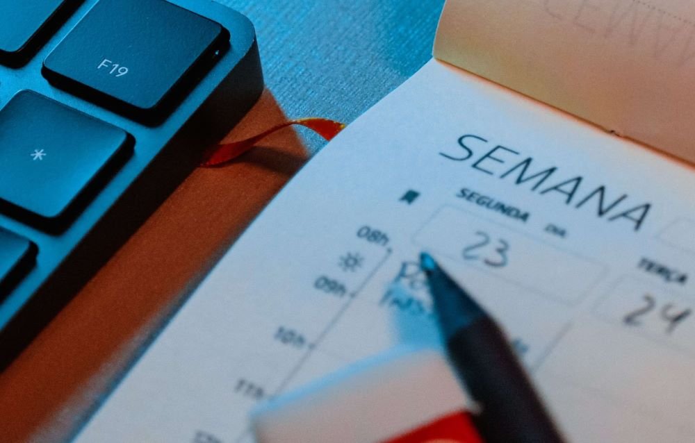Agenda semanal aberta com uma caneta sobre a página, ao lado de um teclado.