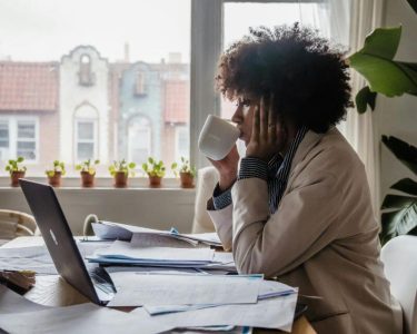 Mulher sentada à mesa de trabalho em casa, observando documentos e contas espalhados enquanto toma café e analisa suas finanças no computador.